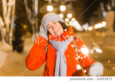 Young beautiful woman in knitted hat and scarf standing in the city with bengal light, sparkler. Concept celebration and christmas. 83645673
