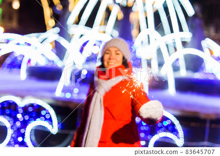 Young woman in knitted hat and scarf standing in the city with bengal light, sparkler. Concept celebration and christmas. Young woman in knitted hat and scarf standing in the city with bengal light, sparkler. Concept celebration and christmas. 83645707