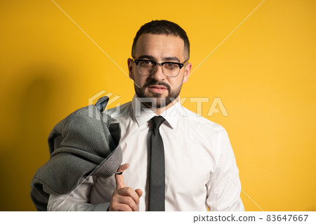 Looking serious business man with trendy beard dressed in greyish suite posing holding his jacket on his shoulder hanging it behind looking isolated on yellow background 83647667