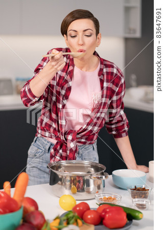 Young housewife with a short hairstyle testing a food while prepares dinner for family in the kitchen. Healthy food at home. Healthy food leaving - vegan concept. Horizontal image 83647691