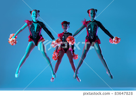 Full-length portrait of three girls, cheerleaders training wearing colorful costumes with pom-pons isolated over blue background in neon lights Full-length portrait of three girls, cheerleaders training wearing colorful costumes with pom-pons isolated over blue background in neon lights 83647828