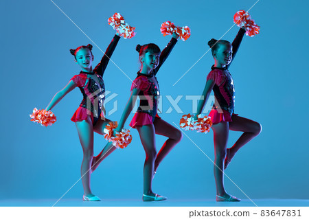 Full-length portrait of three girls, cheerleaders training wearing colorful costumes with pom-pons isolated over blue background in neon lights Full-length portrait of three girls, cheerleaders training wearing colorful costumes with pom-pons isolated over blue background in neon lights 83647831