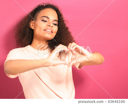 Sending love with gesture of a tender looking beautiful young African American woman looking positively at camera wearing peachy t-shirt isolated on pink background. Beauty concept Sending love with gesture of a tender looking beautiful young African American woman looking positively at camera wearing peachy t-shirt isolated on pink background. Beauty concept 83648100