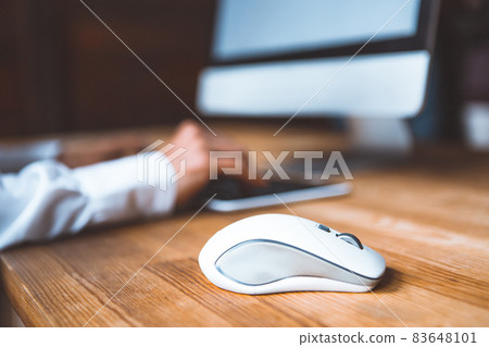 Close-up of a computer mouse on a table against the background of a working girl at a computer dressed in a white shirt. Selected focus on computer mouse on the wooden table Close-up of a computer mouse on a table against the background of a working girl at a computer dressed in a white shirt. Selected focus on computer mouse on the wooden table 83648101