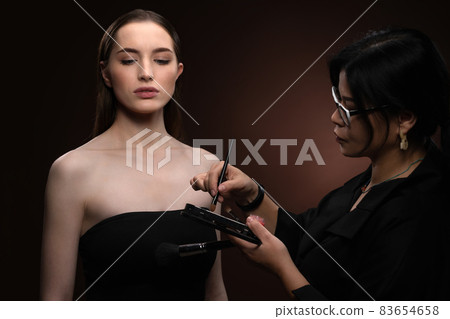 Beauty makeup artist working on a fashion model pretty face isolated on black background. Beautiful model at make up artist. Make-up artist, applying eye shadow to the model Beauty makeup artist working on a fashion model pretty face isolated on black background. Beautiful model at make up artist. Make-up artist, applying eye shadow to the model 83654658
