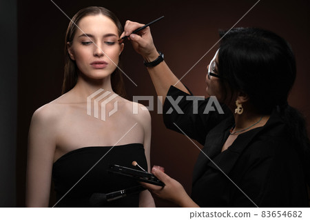 Beautiful model at MUAH artist. Beauty makeup artist working on a fashion model face isolated on black background. Make-up artist, applying eye shadow to the model 83654682