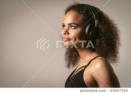 Pretty hipster girl African-American listening music in black earphones wearing black top isolated on grey background, emotionally move. Concept of emotions, facial expression 83657164