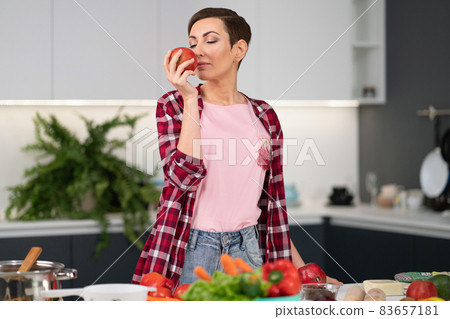 Smelling fresh apple holding it in a hand housewife wearing plaid shirt with a short hairstyle while cooking apple pie standing at the kitchen. Healthy food at home. Healthy food leaving concept Smelling fresh apple holding it in a hand housewife wearing plaid shirt with a short hairstyle while cooking apple pie standing at the kitchen. Healthy food at home. Healthy food leaving concept 83657181