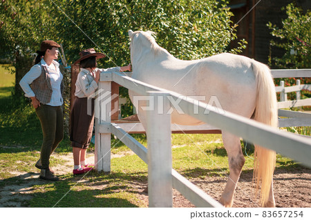 Happy farm living. Mother and daughter in vintage clothes stand near the paddock with a beautiful white horse. The girl is stroking a horse. Family at the ranch Happy farm living. Mother and daughter in vintage clothes stand near the paddock with a beautiful white horse. The girl is stroking a horse. Family at the ranch 83657254