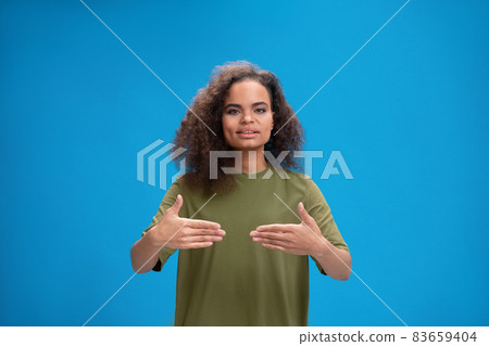 Pretty african american girl saying HELLO using her hands to communicate. Curly haired young woman showing hello sign talking on camera isolated on blue background Pretty african american girl saying HELLO using her hands to communicate. Curly haired young woman showing hello sign talking on camera isolated on blue background 83659404