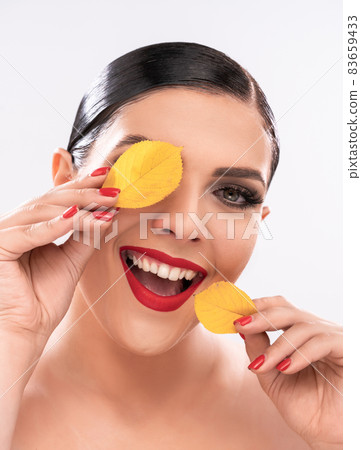 Toothy smiling Caucasian woman holds yellow leaves near face like patches. Beauty concept. Isolated on white background. Close up shot Toothy smiling Caucasian woman holds yellow leaves near face like patches. Beauty concept. Isolated on white background. Close up shot 83659433