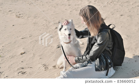 young woman wearing jacket and dress sitting on a sand next to her dog. white yakutian laika and owner spending time and relaxing together on a beach 83660052
