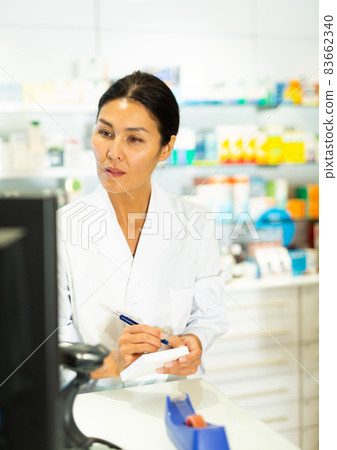 Medicine, profession and healthcare concept - female doctor or pharmacist in white coat with notebook and pen writing medical report over pharmacy 83662340