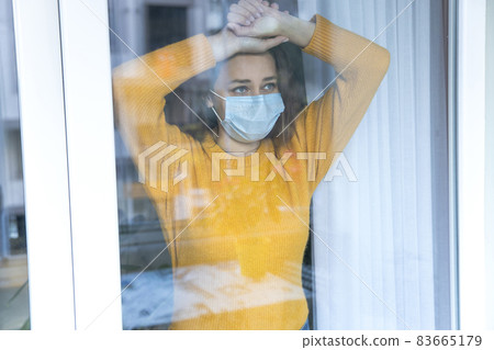 Young woman in quarantine wearing a mask and looking through the window 83665179