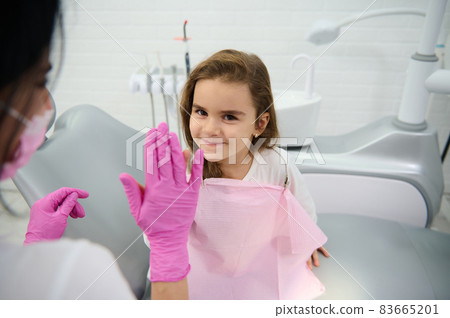 Adorable little child sitting in dentist's chair after medical check-up, smiles looking at camera and gives high five to her treating pediatric dentist. Beautiful girl visiting children dentist Adorable little child sitting in dentist's chair after medical check-up, smiles looking at camera and gives high five to her treating pediatric dentist. Beautiful girl visiting children dentist 83665201
