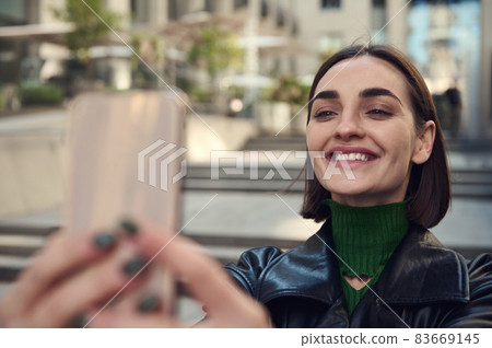 Close-up lifestyle portrait of beautiful funny young brunette with stylish short hair, holding smartphone in outstretched hands and smiling toothy smile taking selfie while standing outdoors in city 83669145