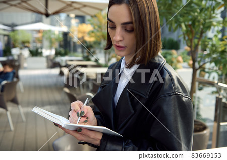Confident portrait of concentrated young business woman, copywriter, journalist, writing on white notepad while sitting outdoors on urban public background. Close-up Confident portrait of concentrated young business woman, copywriter, journalist, writing on white notepad while sitting outdoors on urban public background. Close-up 83669153