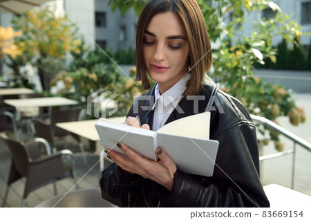 Confident portrait of focused young woman writing on white notepad while sitting outdoors on urban public background 83669154