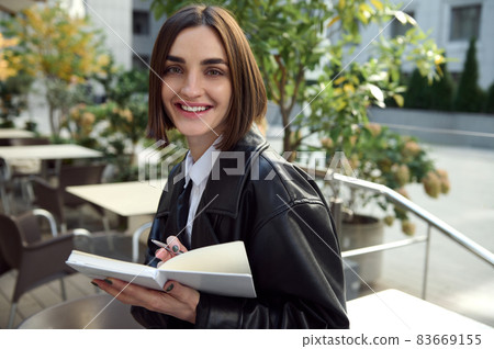 Portrait of attractive young copywriter, freelancer woman with short hair, cute smiling looking at camera, holding a notepad and writing texts enjoying outside warm weather Portrait of attractive young copywriter, freelancer woman with short hair, cute smiling looking at camera, holding a notepad and writing texts enjoying outside warm weather 83669155