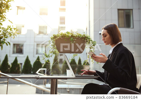 Concentrated freelance woman in business casual clothes holds a mobile phone and types on laptop keyboard, works remotely sitting on a summer terrace of a street cafe with high buildings on background 83669184