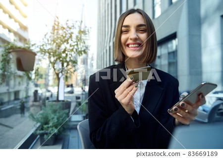 Online shopping, remote work, business and finance concept. Beautiful woman, entrepreneur, with smartphone and gold credit card , smiles toothy smile standing against urban modern buildings background Online shopping, remote work, business and finance concept. Beautiful woman, entrepreneur, with smartphone and gold credit card , smiles toothy smile standing against urban modern buildings background 83669189