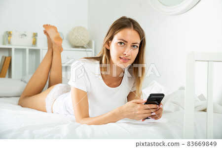 Young woman in lace lingerie lying in bed with mobile phone in hands in morning sunbeams 83669849