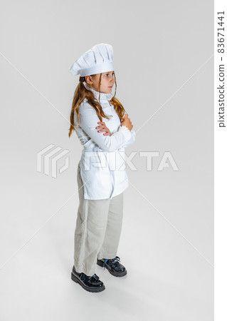 Full-length portrait of little girl in white cook uniform and huge chef's hat posing isolated on white studio background. Full-length portrait of little girl in white cook uniform and huge chef's hat posing isolated on white studio background. 83671441