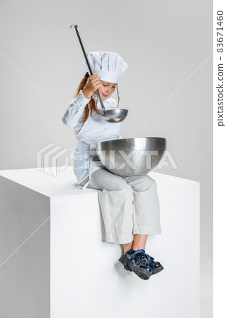 Photography of little girl, student in white cook uniform and huge chef's hat sitting on huge box isolated on white studio background. 83671460