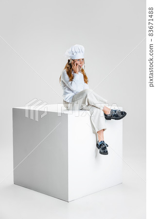Photography of little girl, student in white cook uniform and huge chef's hat sitting on huge box isolated on white studio background. Photography of little girl, student in white cook uniform and huge chef's hat sitting on huge box isolated on white studio background. 83671488