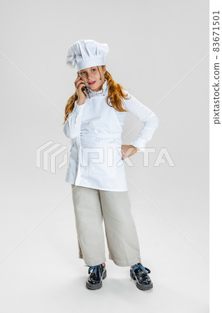 One little cute girl in white cook uniform and huge chef's hat posing isolated on white studio background. 83671501