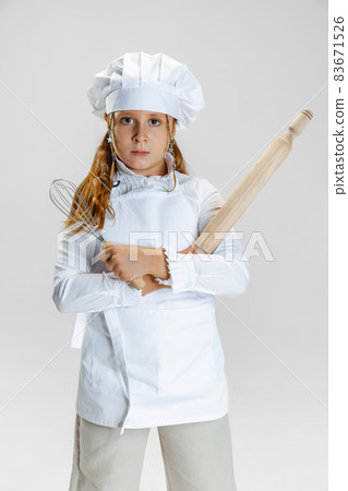 One little cute girl in white cook uniform and huge chef's hat posing isolated on white studio background. One little cute girl in white cook uniform and huge chef's hat posing isolated on white studio background. 83671526