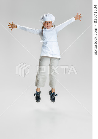Dynamic image of little cute girl in white cook uniform and huge chef's hat jumping isolated on white studio background. Dynamic image of little cute girl in white cook uniform and huge chef's hat jumping isolated on white studio background. 83671534