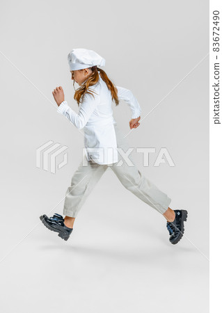 Little Caucasian girl in white cook uniform and huge chef's hat running isolated on white studio background. 83672490
