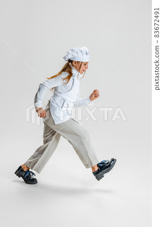 Little Caucasian girl in white cook uniform and huge chef's hat going isolated on white studio background. 83672491