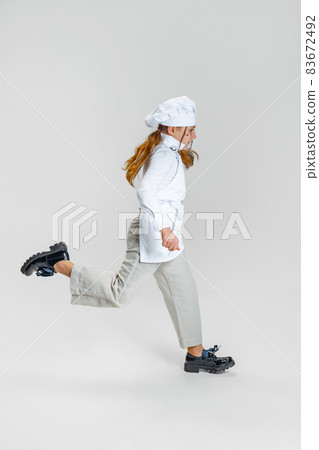 Little Caucasian girl in white cook uniform and huge chef's hat running isolated on white studio background. 83672492