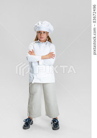 Portrait of little cute girl in white cook uniform and huge chef's hat standing with arms crossed isolated on white studio background. 83672496