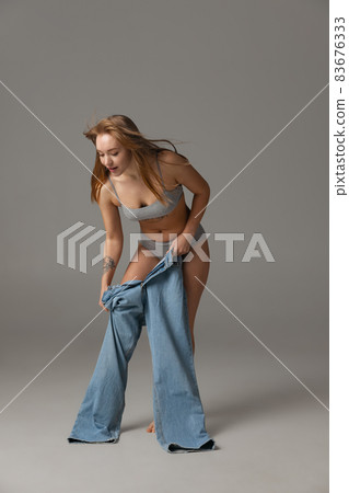Young beautiful red-headed woman putting blue jeans jumpersuit on isolated over gray studio background. Weight Loss concept. 83676333