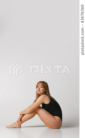 Young beautiful red-headed girl in black swimsuit sitting on floor isolated over gray studio background. Natural beauty concept. Young beautiful red-headed girl in black swimsuit sitting on floor isolated over gray studio background. Natural beauty concept. 83676360