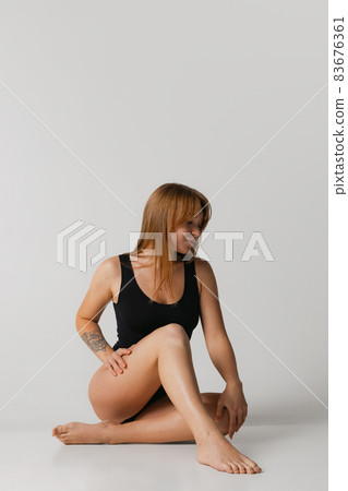 Young beautiful red-headed girl in black swimsuit sitting on floor isolated over gray studio background. Natural beauty concept. Young beautiful red-headed girl in black swimsuit sitting on floor isolated over gray studio background. Natural beauty concept. 83676361