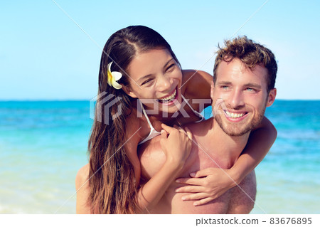Beach vacation happy couple in love having fun lifestyle. Asian woman piggybacking hugging man in summer vacation. Smiling young people on travel holidays. 83676895