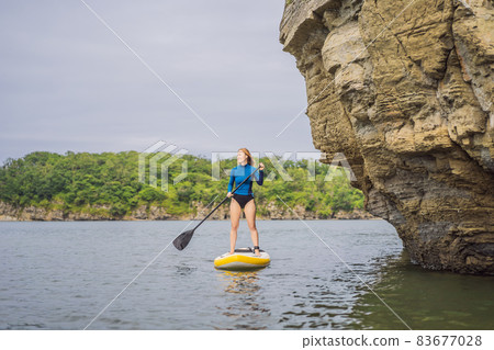 Young women Having Fun Stand Up Paddling in the sea. SUP. Red hair girl Training on Paddle Board near the rocks Young women Having Fun Stand Up Paddling in the sea. SUP. Red hair girl Training on Paddle Board near the rocks 83677028