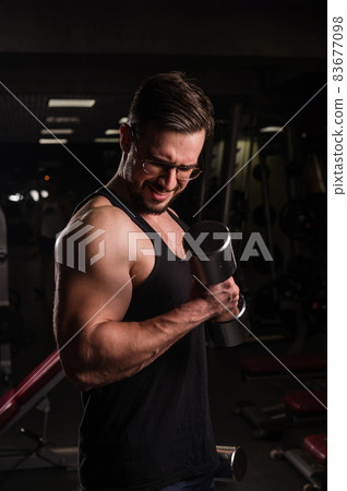A man with an effort does an exercise for biceps and triceps with a dumbbell. Embossed bodybuilder. Muscular guy with glasses in the gym. A man with an effort does an exercise for biceps and triceps with a dumbbell. Embossed bodybuilder. Muscular guy with glasses in the gym. 83677098