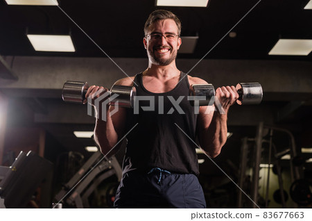 A man with an effort does an exercise for biceps and triceps with a dumbbell. Embossed bodybuilder smiles. Muscular guy with glasses in the gym. 83677663