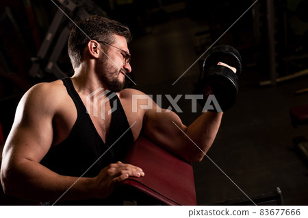 A handsome man in glasses with a beard is doing an exercise for biceps with a barbell. The young guy is engaged in bodybuilding. The trainer in the gym smiles with muscular arms. A handsome man in glasses with a beard is doing an exercise for biceps with a barbell. The young guy is engaged in bodybuilding. The trainer in the gym smiles with muscular arms. 83677666