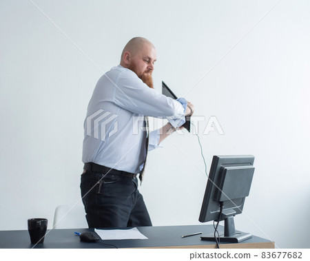 Angry bald man with a red beard in the office in a business suit crashes a computer. Manager with a nervous breakdown breaks the keyboard on the monitor. 83677682