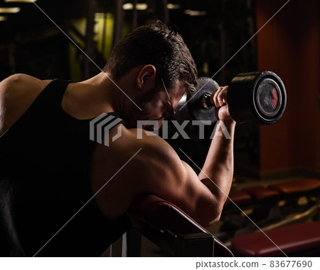 A handsome man with glasses doing an exercise for biceps with a barbell. The guy is engaged in bodybuilding. Trainer in the gym with muscular arms. A handsome man with glasses doing an exercise for biceps with a barbell. The guy is engaged in bodybuilding. Trainer in the gym with muscular arms. 83677690