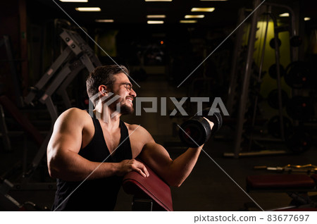 A handsome man in glasses with a beard is doing an exercise for biceps with a barbell. The young guy is engaged in bodybuilding. The trainer in the gym smiles with muscular arms. 83677697