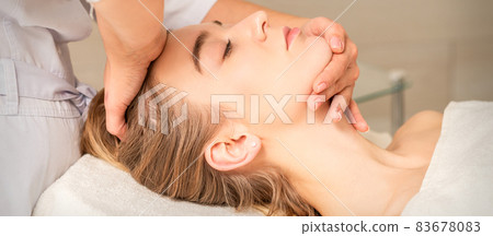 Cosmetologist hands doing facial massage on forehead and chin of young female at spa salon. 83678083