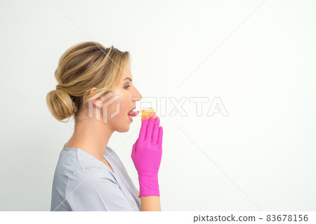 Young beautician licks liquid wax for depilation holding in gloved hands standing on white background. Cosmetologist tastes the sugar paste to taste. Young beautician licks liquid wax for depilation holding in gloved hands standing on white background. Cosmetologist tastes the sugar paste to taste. 83678156