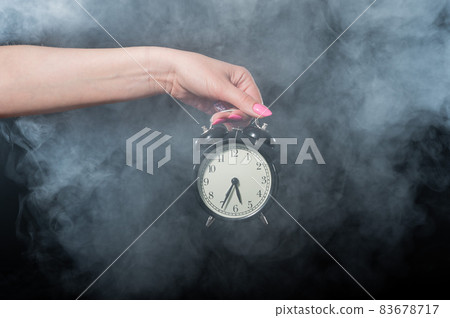 A woman holds an alarm clock in a studio full of smoke. White fog enveloped a round retro mechanical watch. A woman holds an alarm clock in a studio full of smoke. White fog enveloped a round retro mechanical watch. 83678717